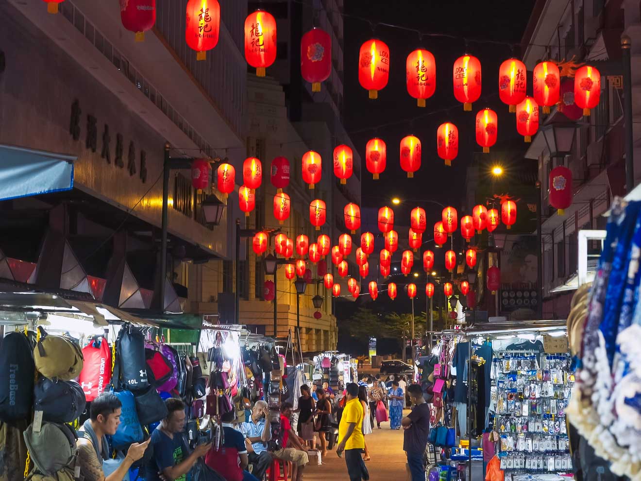 Bustling night market scene under a canopy of glowing red lanterns with vendors selling various goods and people milling about in a lively street in Chinatown