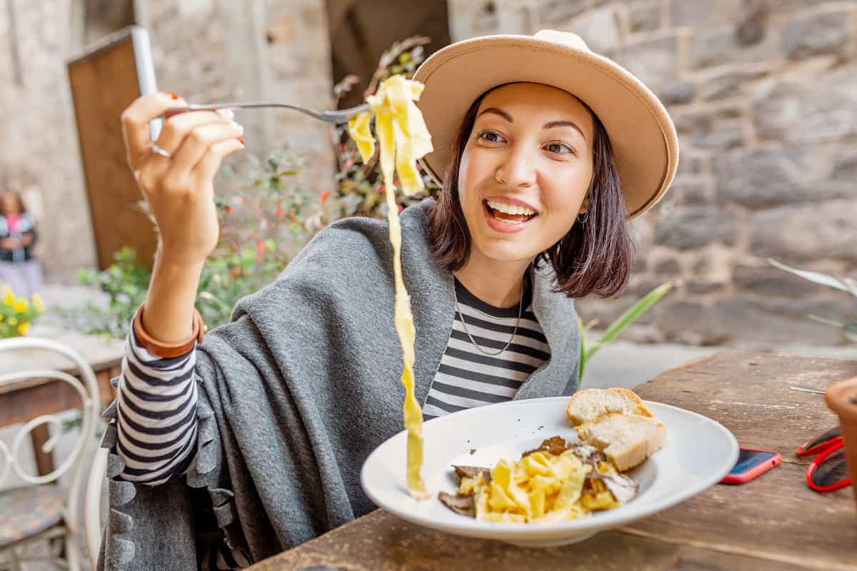 A happy asian woman wearing a hat holds up a string of pasta with her fork in a roman restaurant. Rome food tour