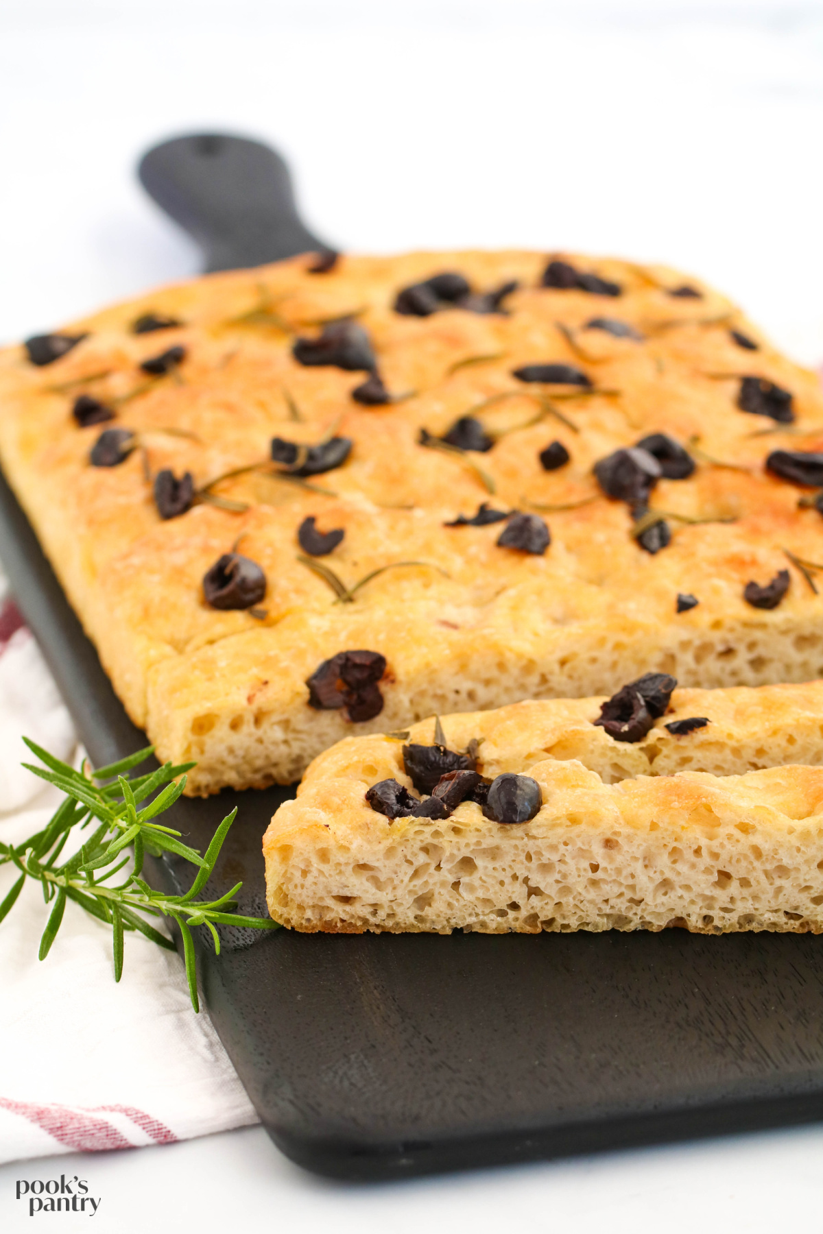 Rosemary and black olive focaccia on black board with rosemary sprigs on the side.