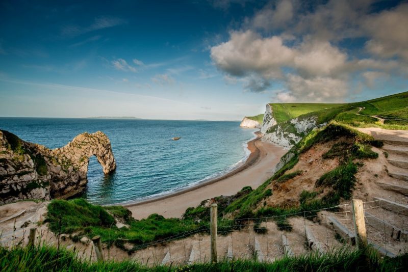 view of Durdle Door a stone archway over the sea next to a curved bay with blue sea - seen from a grassy cliff top with the south west coast path leading past